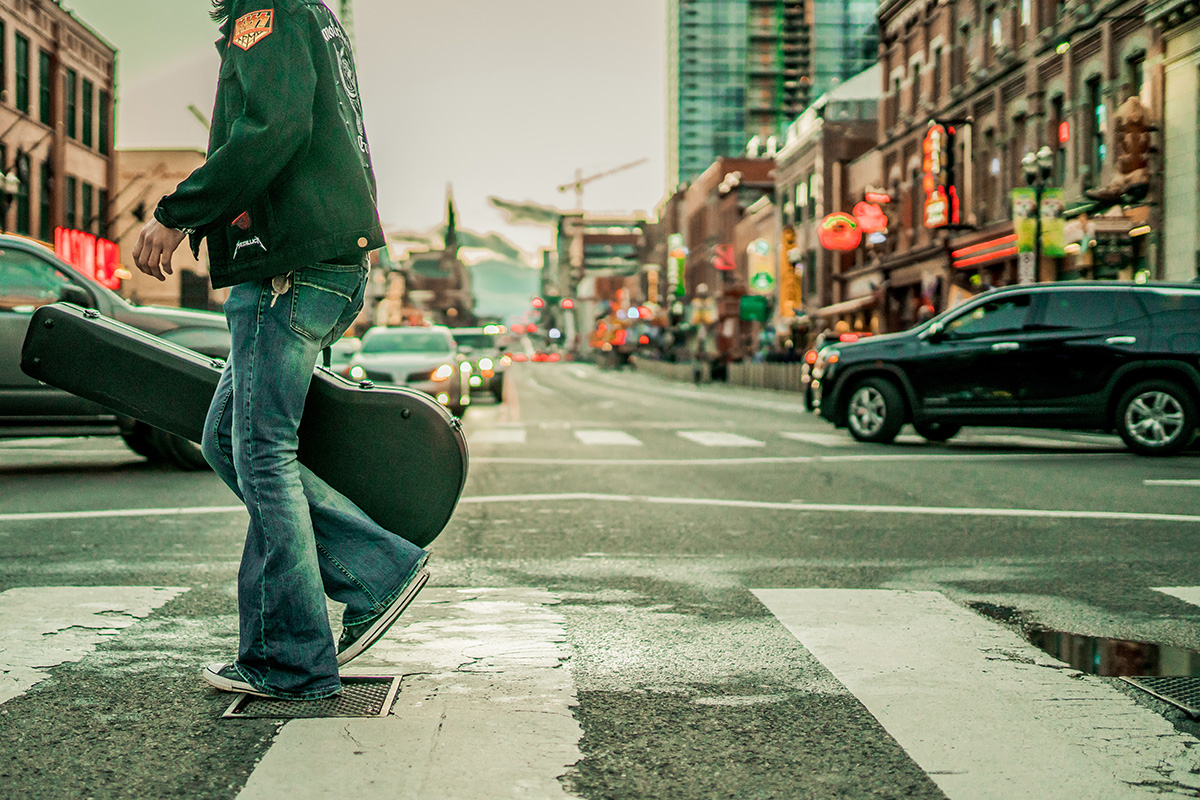 a person wearing bell bottoms and a jacket with band logos crosses an intersection carrying a guitar case